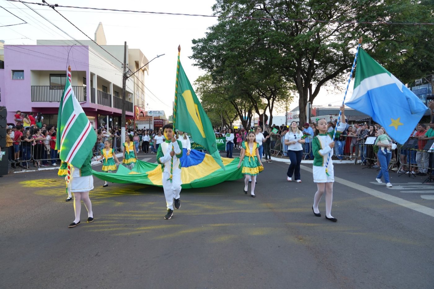 Estudantes de uniforme verde, branco e amarelo desfilam na avenida carregando uma grande bandeira do Brasil e a bandeira de Mato Grosso do Sul, com o público acompanhando atrás das grades.